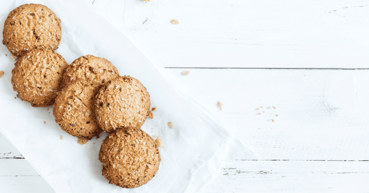 Five oatmeal cookies are arranged on white parchment paper atop a white wooden surface, with a few crumbs scattered nearby—a simple yet mindful treat for anyone on the journey of overcoming binge eating.