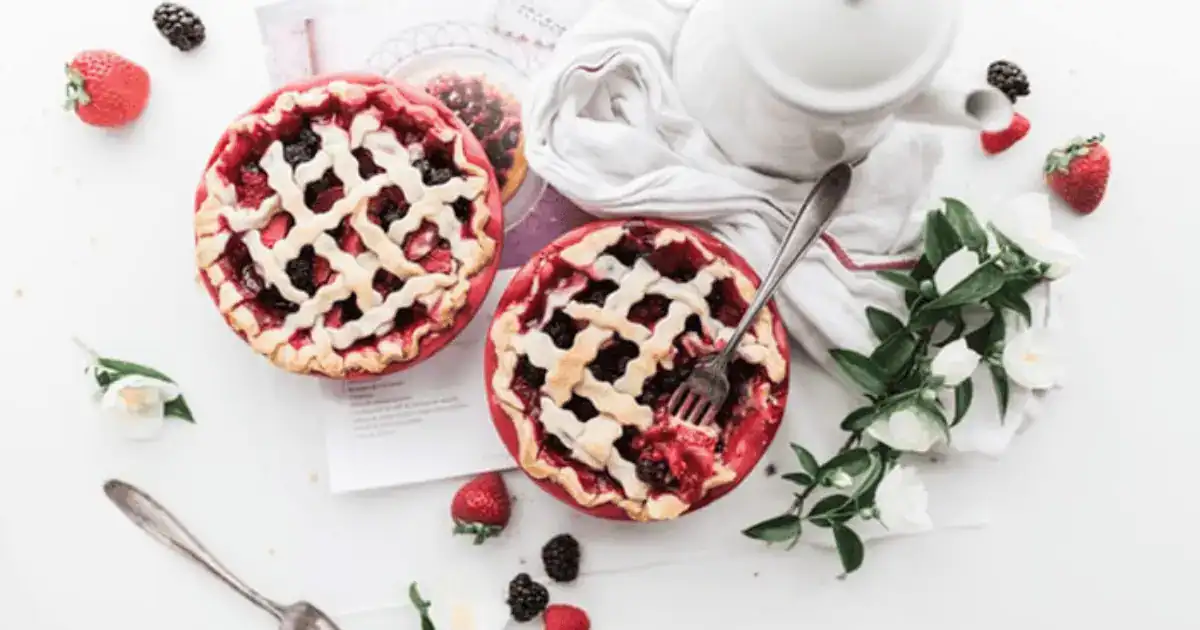 Two berry pies with lattice crusts sit on a table, surrounded by strawberries, blackberries, a white teapot, flowers, a spoon, and a white cloth napkin. A fork rests on one of the pies, capturing the comforting scene that evokes feelings of being emotional about eating—where each bite offers solace and sweetness in equal measure.