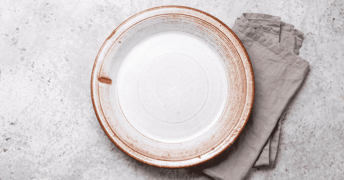 A large white ceramic plate with brown edging sits on a light gray textured surface, next to a neatly folded beige cloth napkin—perfect for illustrating topics like ARFID explained, where food presentation and mealtime environments play an important role.