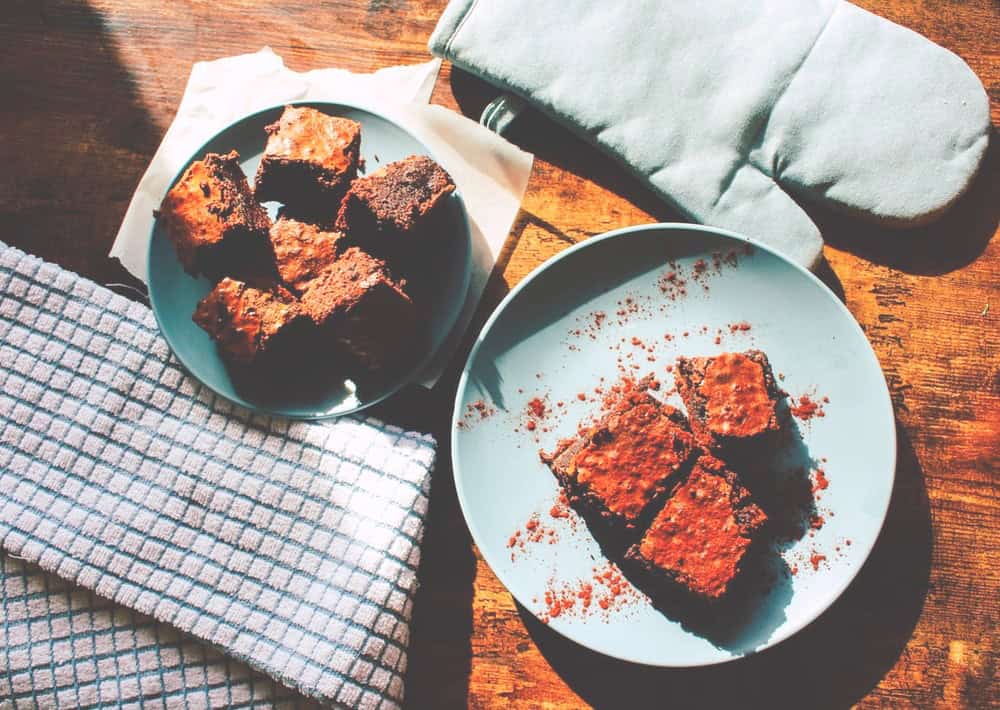 Two blue plates with chocolate brownies on a wooden table, next to a gray oven mitt and a white dish towel with a blue stripe grid pattern. Sunlight casts strong shadows across the scene, creating an inviting atmosphere that hints at Why You Eat When You’re Not Hungry—sometimes the mood and beauty of food are simply irresistible.