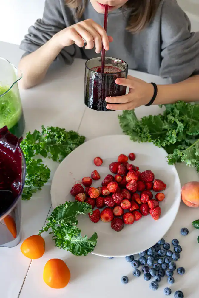 A person drinks a dark smoothie at a table with fresh kale, strawberries, blueberries, apricots, and a peach on a white plate and surface.